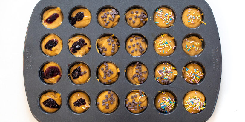 unbaked mini flourless peanut butter muffins in muffin tin topped with sprinkles, chocolate chips and jelly