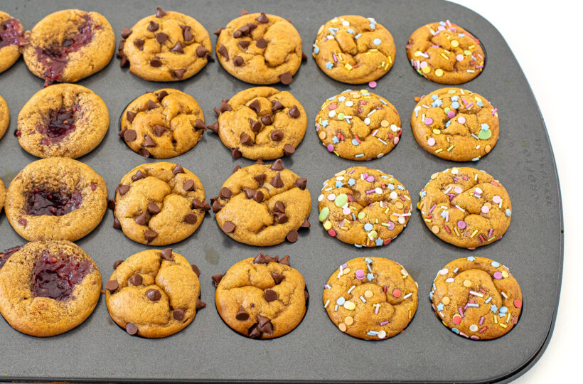 Mini peanut butter muffins with a variety of toppings in a muffin tin after being baked. 