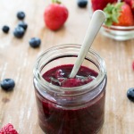 Mixed berry compote in glass jar with spoon on wooden table next to fresh berries.