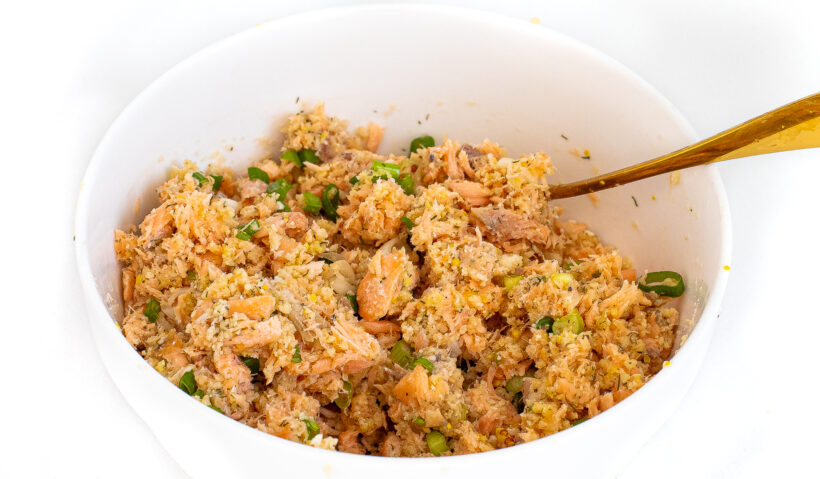 A fork stirring the ingredients for salmon cakes in a white mixing bowl.
