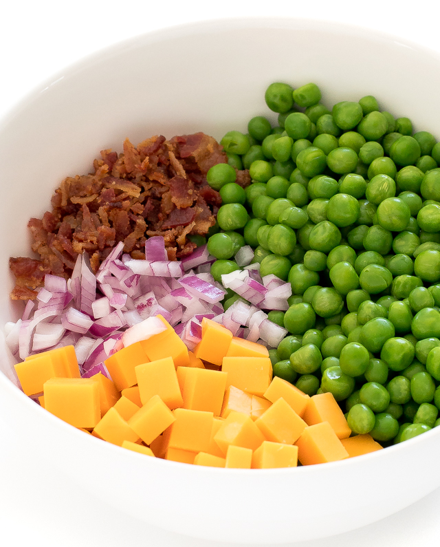 cubed cheese, red onion, crumbled bacon and peas in large white bowl