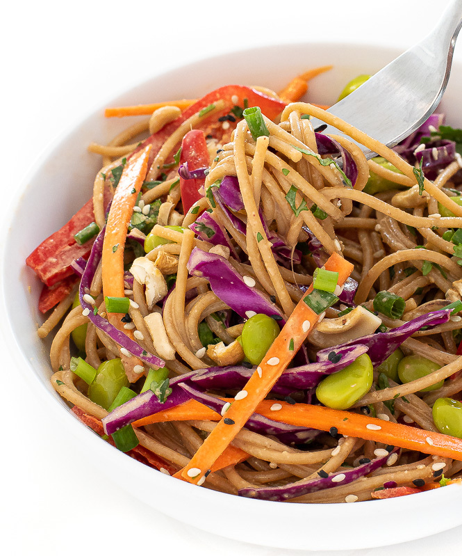 Rainbow peanut noodles in a white bowl with a silver fork spinning the noodles.