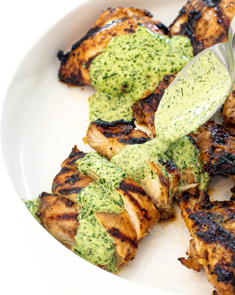 Grilled chicken on a white plate with a spoon pouring green sauce on top. 