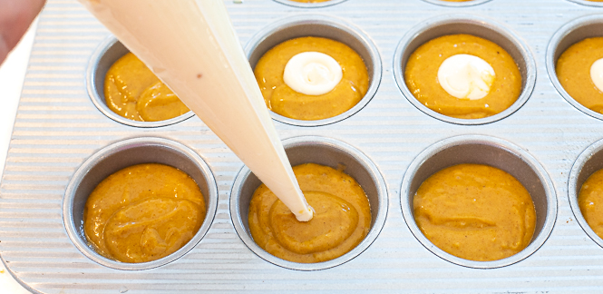 Show of muffin tin with muffin batter inside the cavities and cream cheese being piped into them.