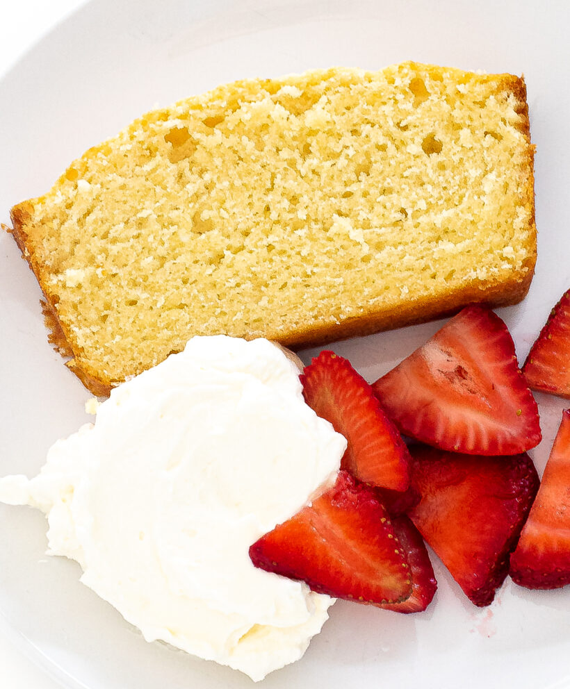 Slice of sour cream pound cake next to a dollop of whipped cream and some fresh strawberries. 