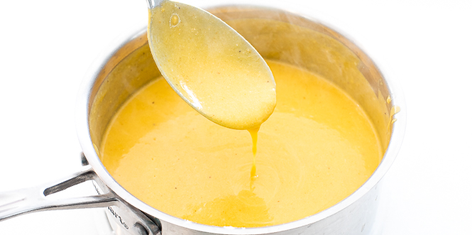 Mac and cheese sauce in a silver pan with a spoon being dipped in the sauce.