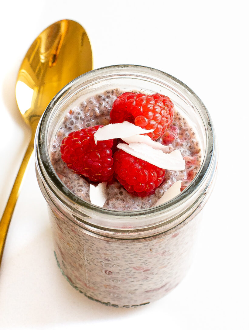 Raspberry chia pudding in a jar topped with fresh raspberries and coconut flakes with a spoon next to it.