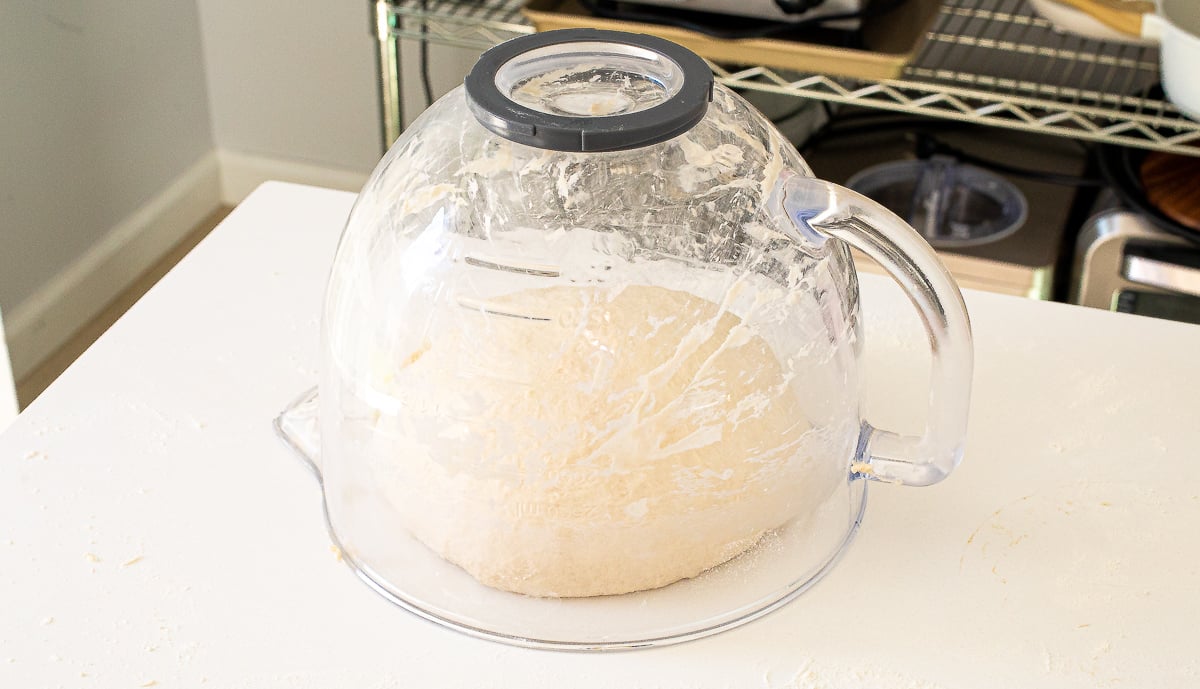 A ball of sourdough sandwich bread dough resting on a white surface, covered by an inverted clear glass mixing bowl.