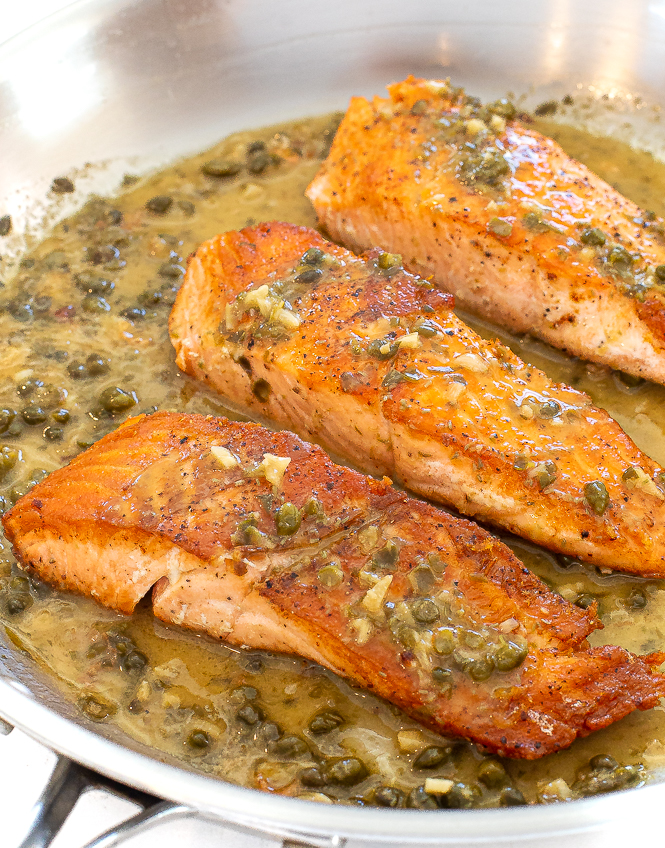 Three salmon filets in a caper sauce in a silver sauté pan.