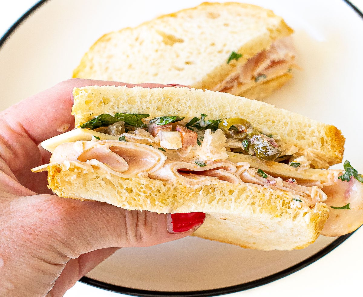 A hand holding a half-sandwich with sliced turkey, cheese, and olives on crusty bread.