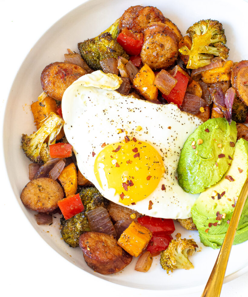 Veggies and sausage served in a bowl topped with a fried egg and avocado slices. 