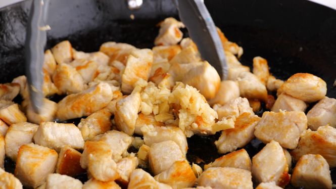 Garlic and ginger added to the cooked chicken in a black pan with tongs.