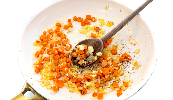 Diced onions and carrots in a white skillet with a wooden spoon.