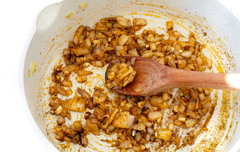 Onions and spices in a skillet with a wooden spoon stirring it.
