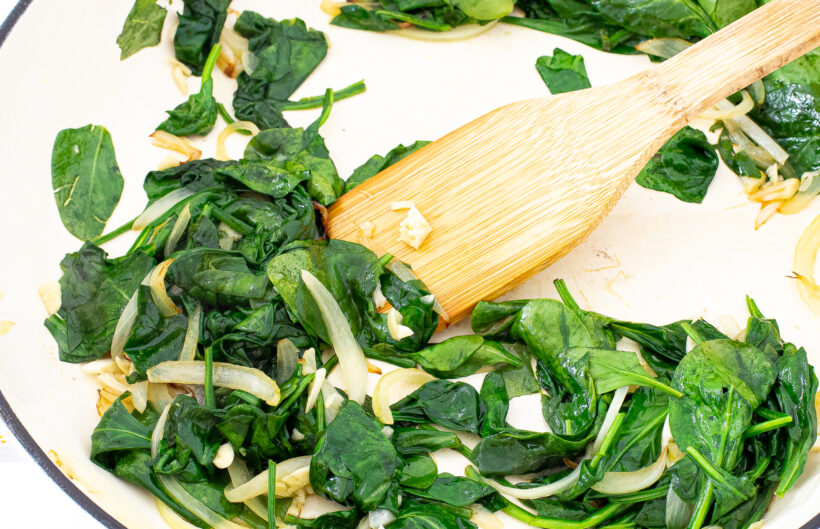 Spinach and onions in a skillet with a wooden spatula stirring it.