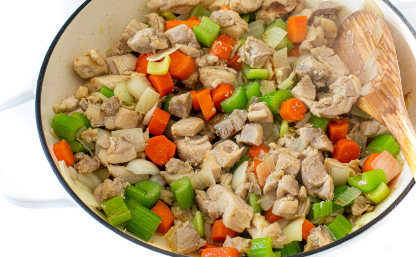 Chunks of chicken, carrots, onion and celery in a white skillet with a wooden spoon stirring it.