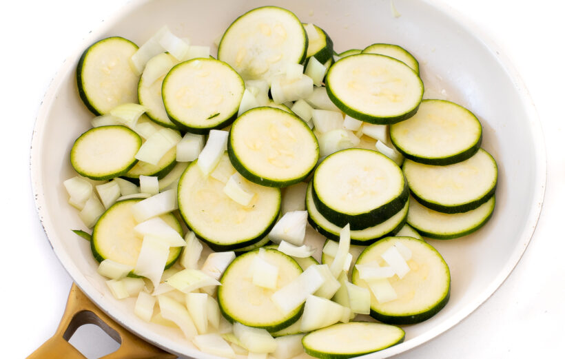 Zucchini slices and diced onion in a white skillet.