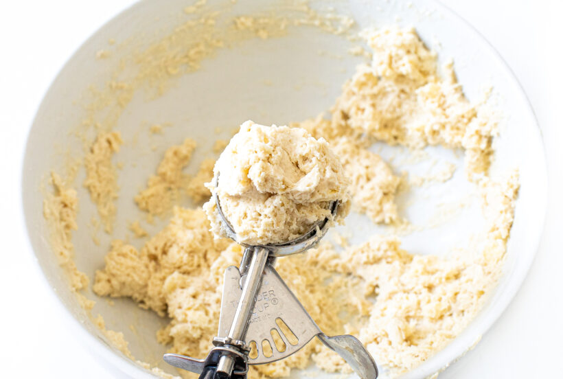 Biscuit dough in a mixing bowl with a cookie scooper scooping up biscuits. 