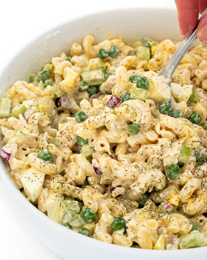 Hand holding spoon filled with macaroni salad with bowl of macaroni salad below it. 