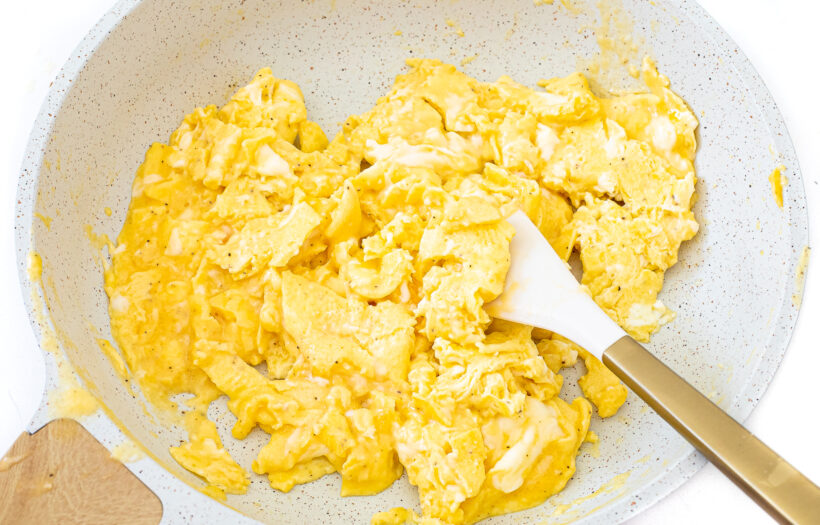 Scrambled eggs in a white frying pan with a white and gold spatula.