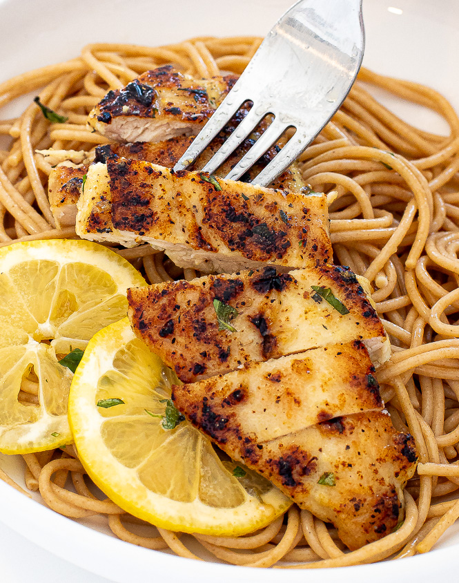 Sliced lemon pepper chicken on a bed of noodles with a silver fork taking a piece.
