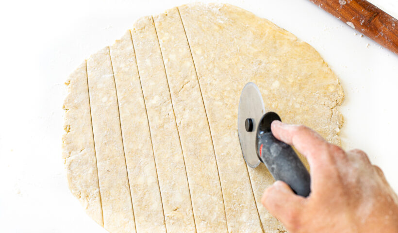 Hand using pizza cutter to cut pie crust dough.