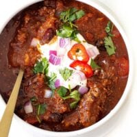 A bowl of Slow Cooker Chili topped with sour cream, chopped red onions, sliced red chili, and fresh cilantro, served with a gold spoon in the bowl.