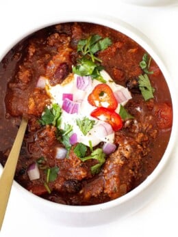 A bowl of Slow Cooker Chili topped with sour cream, chopped red onions, sliced red chili, and fresh cilantro, served with a gold spoon in the bowl.
