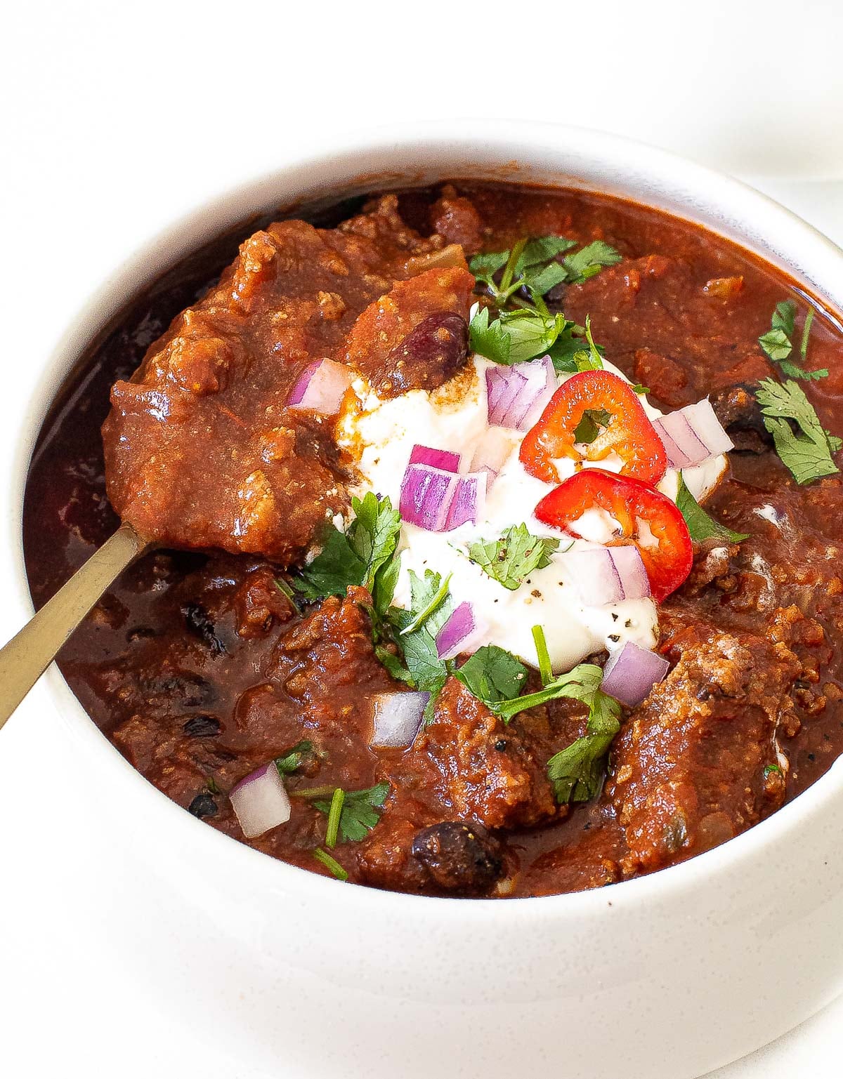 A bowl of Slow Cooker Chili topped with sour cream, chopped red onion, sliced red chili, and cilantro, with a spoon resting inside.
