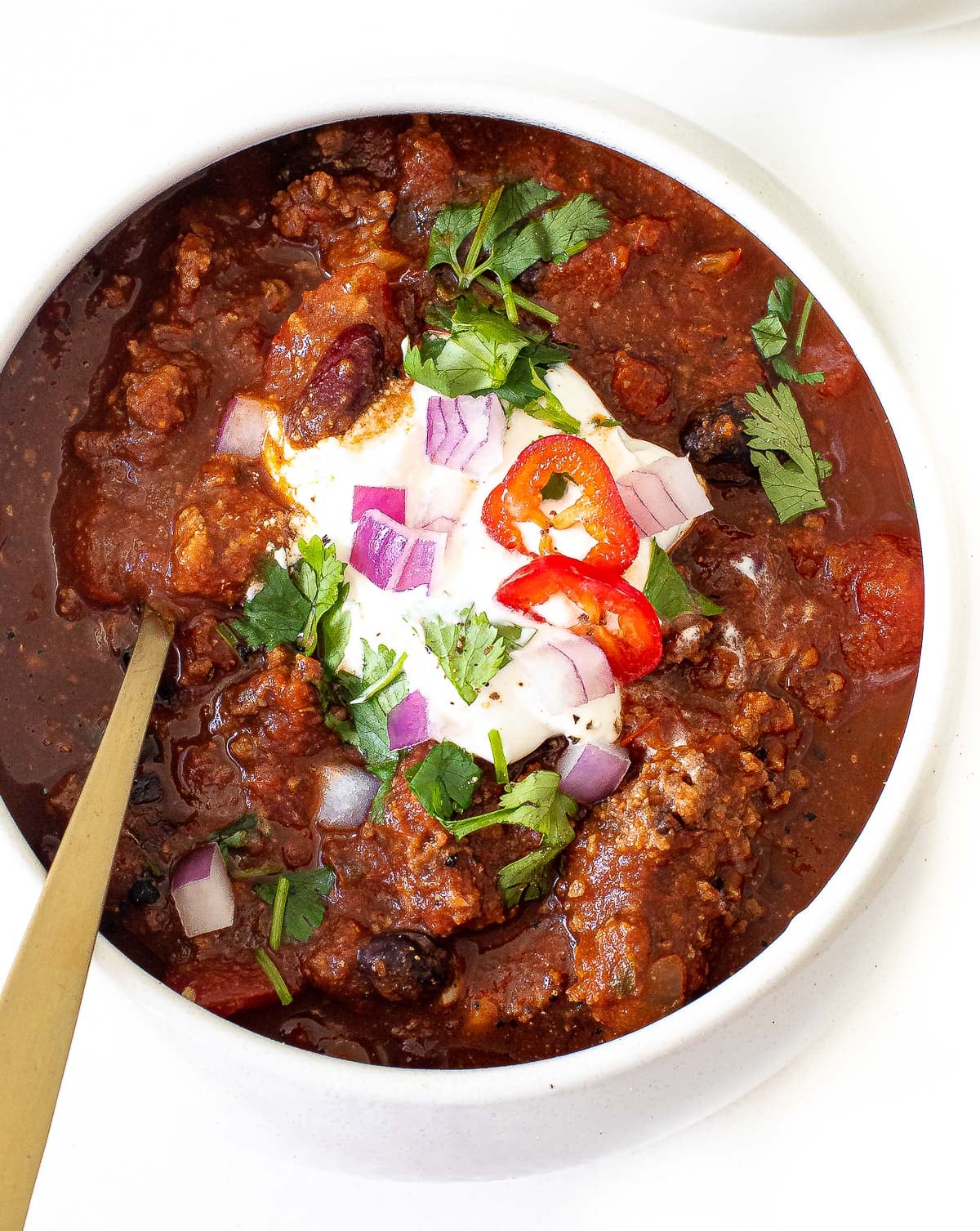 A bowl of Slow Cooker Chili topped with sour cream, chopped red onions, sliced red chili, and fresh cilantro, served with a gold spoon in the bowl.
