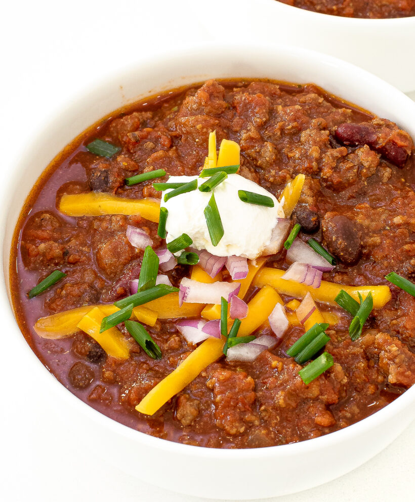beef chili in white bowl with shredded cheddar cheese, sour cream, red onion and green onions