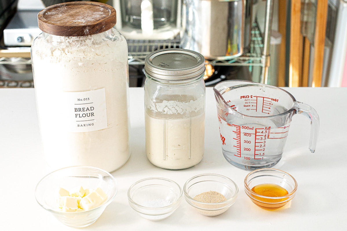 A large jar of bread flour, a jar of sourdough starter, a measuring cup of water, and four small bowls containing butter, salt, yeast, and honey arranged for making sourdough sandwich bread on a white surface.