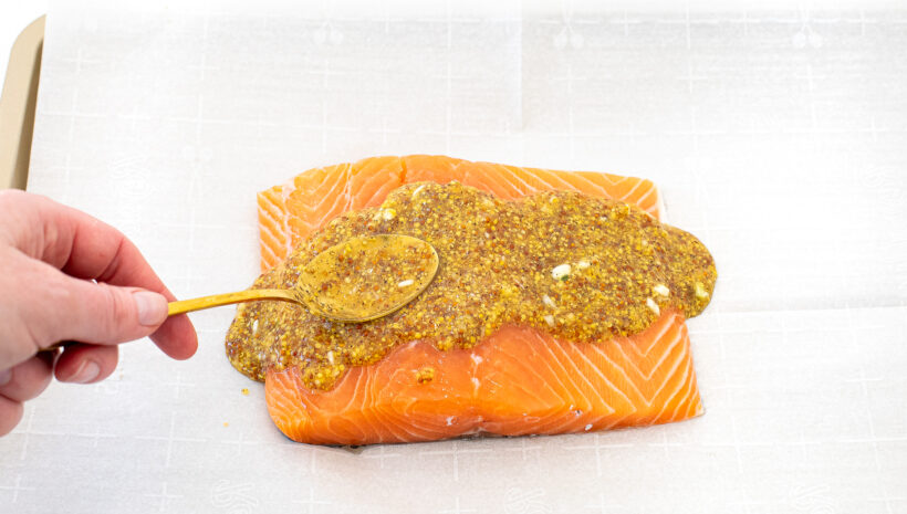 Hand using a spoon to spread the honey mustard sauce on raw salmon. 
