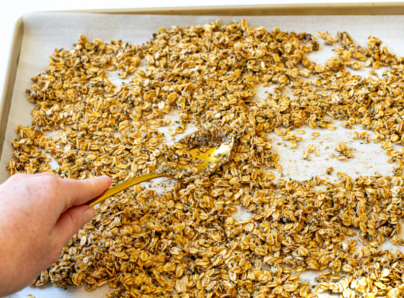 Granola being spread out with a spoon on a baking sheet lined with parchment paper. 