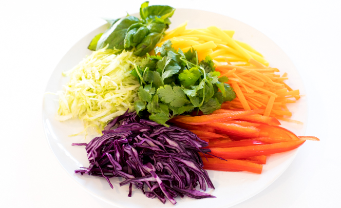 thinly sliced fruits and vegetables like cabbage, bell pepper, carrots, and mango on a white plate with basil and cilantro