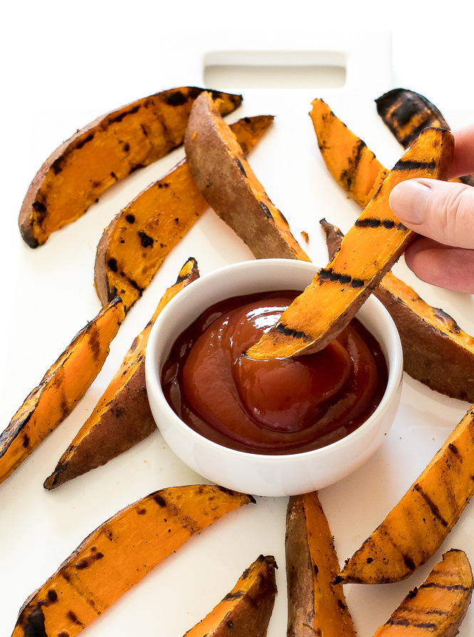 Easy Grilled Sweet Potato Wedges dipped in ketchup Hand dipping a sweet potato wedge into a small bowl of ketchup.