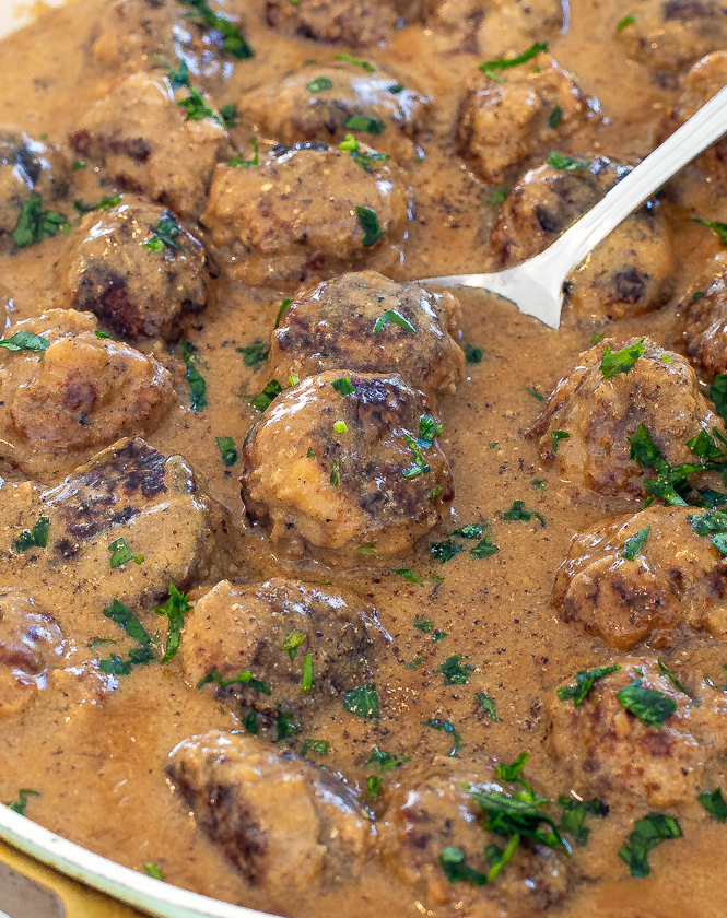Meatballs in a skillet simmering in brown sauce with a metal spoon in the skillet.