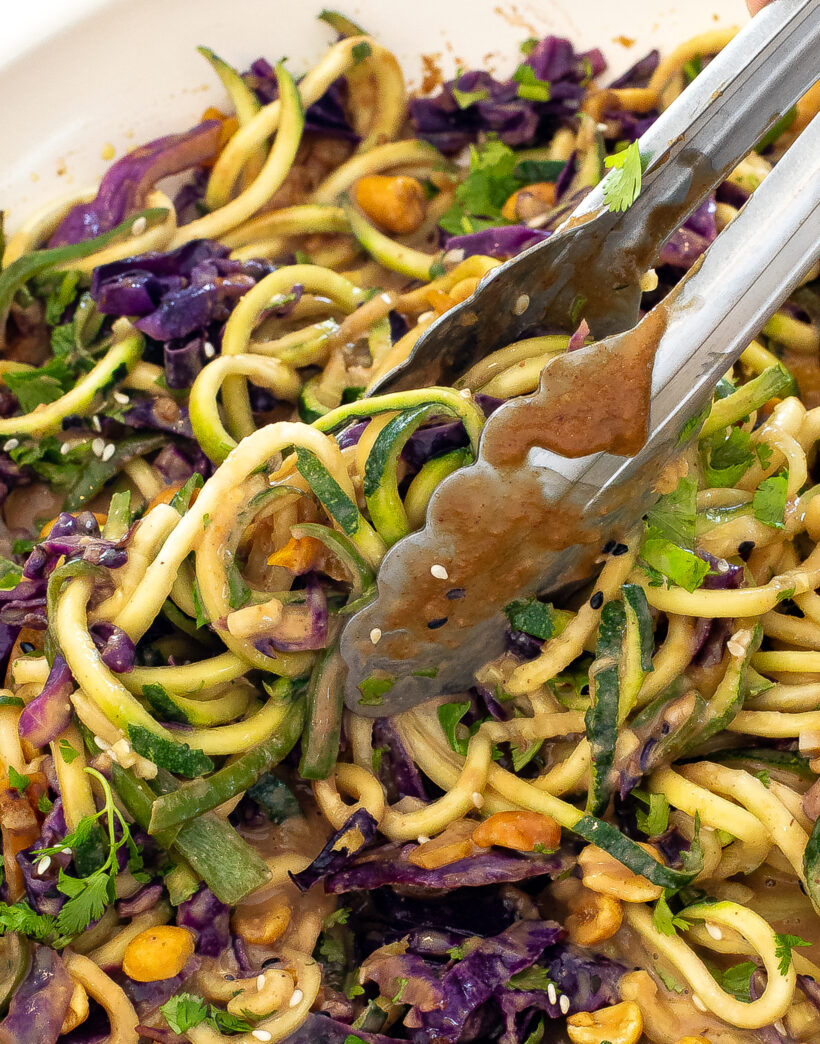 Silver tongs grabbing zucchini peanut noodles. 