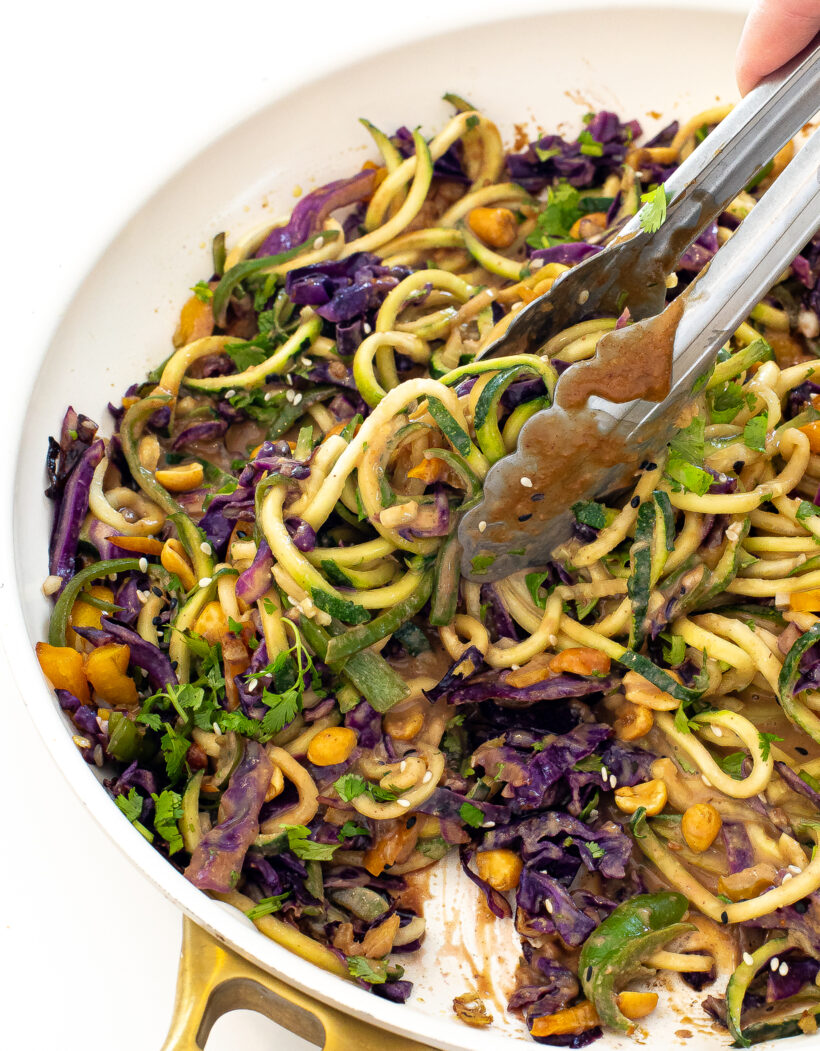 Tongs grabbing a portion of zucchini noodles with peanut sauce. 