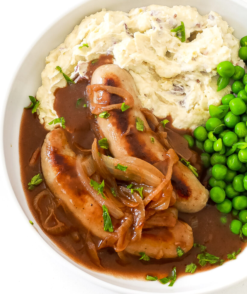Two sausages drenched in onion gravy on a bed of mashed potatoes and a side of peas.
