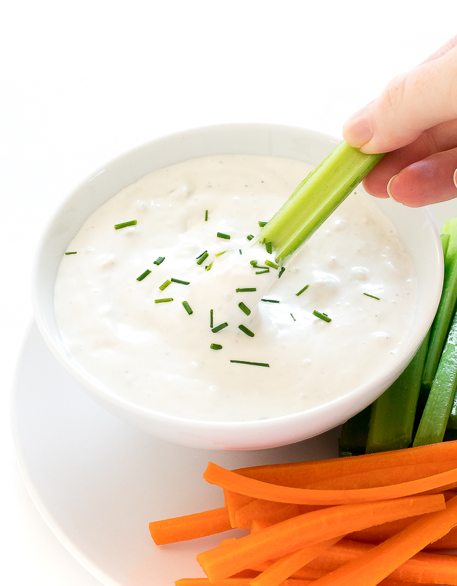 Celery being dipped into Blue Cheese Dressing in a white bowl on a white plate with carrot and celery sticks.
