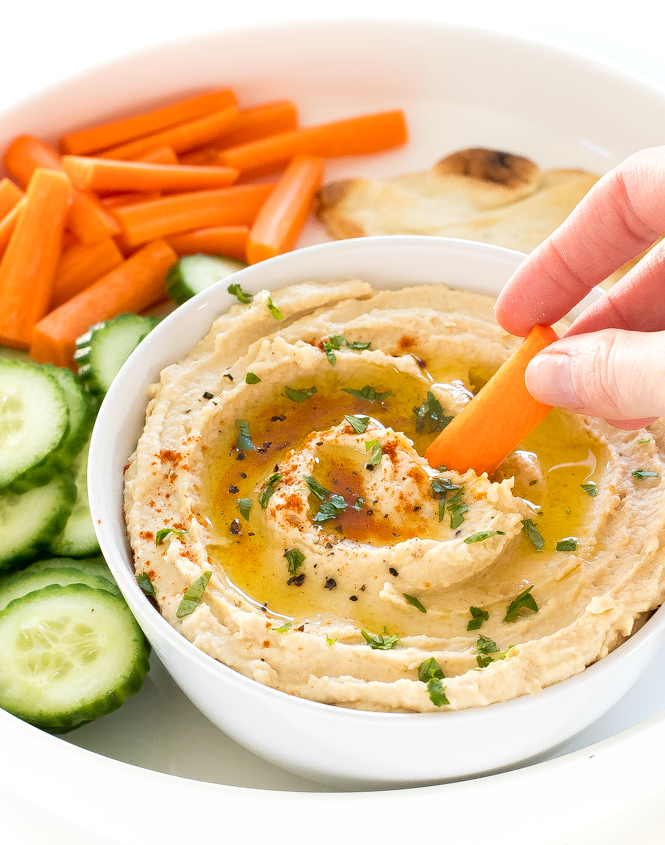 Person dipping baby carrot into bowl of roasted garlic hummus with an olive oil drizzle on top.
