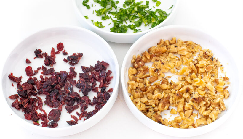 Three white bowls with toppings for the cheeseball: cranberries, walnuts, and parsley.