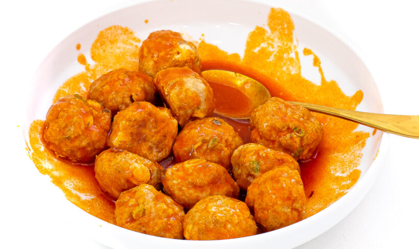 Blue cheese meatballs covered with buffalo sauce in a white serving bowl with a gold spoon.