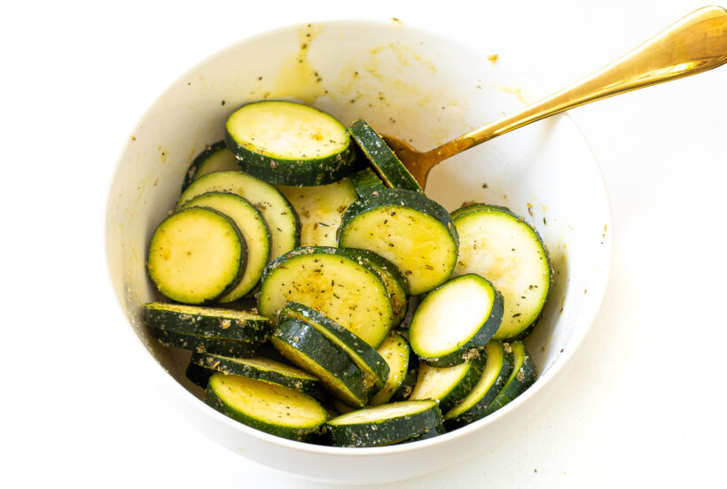 Zucchini slices in a bowl being mixed with olive oil and spices.