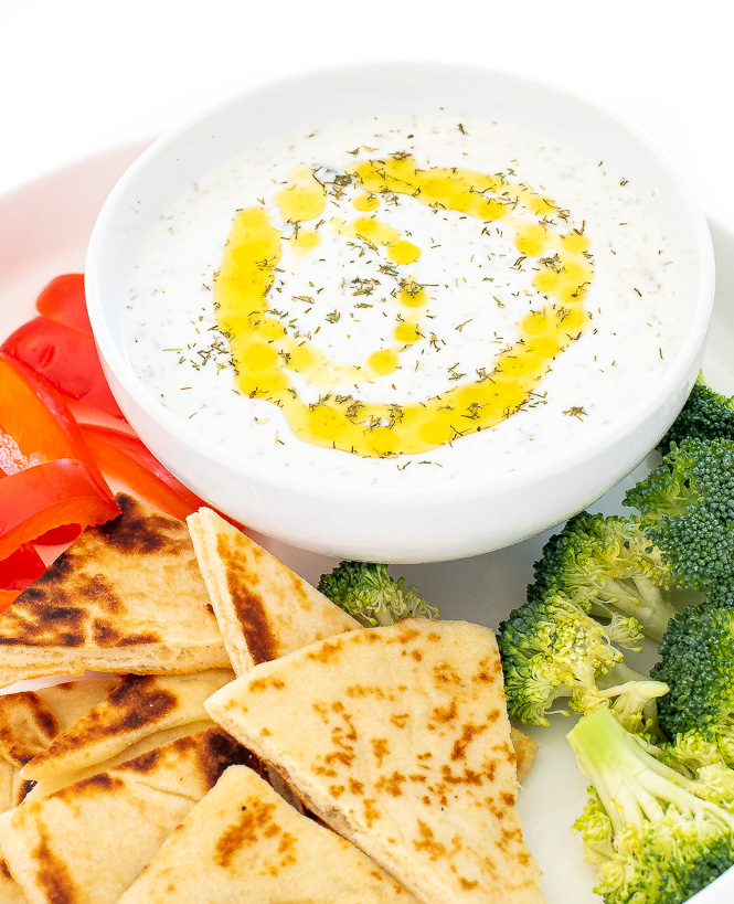 Tzatziki sauce in a white bowl surrounded by veggies and pita for dipping.