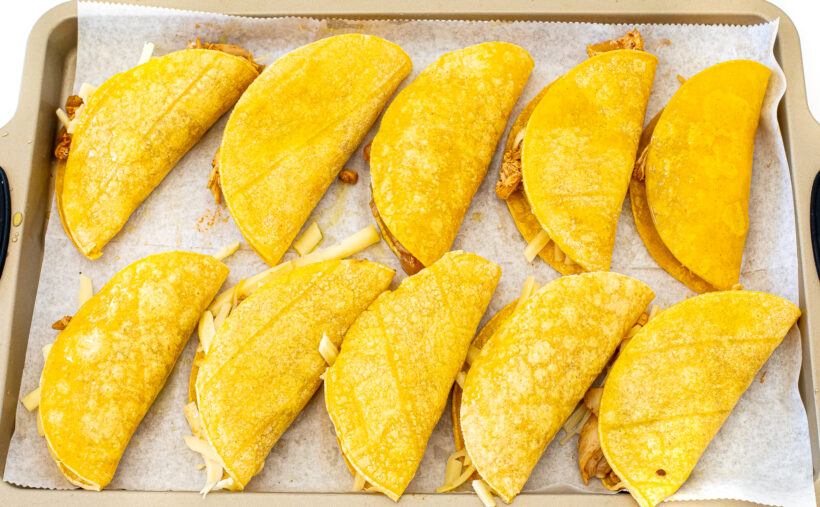 Crispy tacos filled with shredded chicken mixture and placed on a sheet pan.