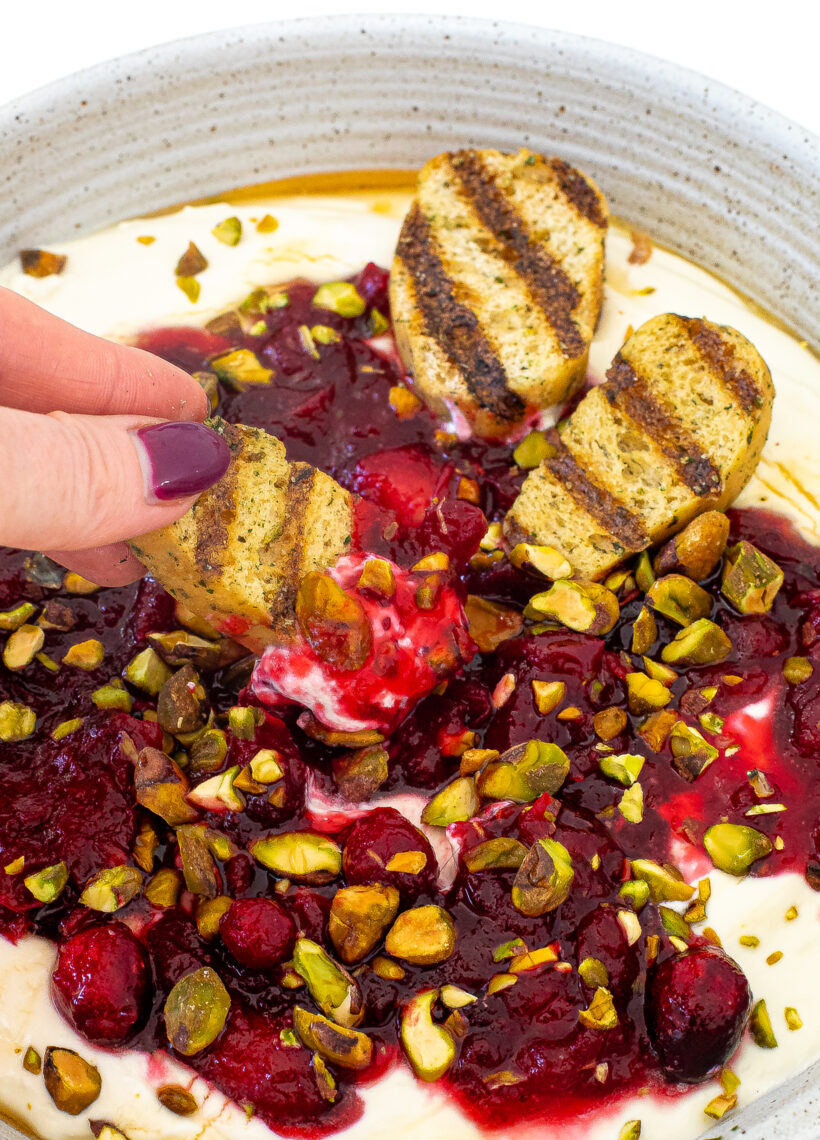 A hand grabbing a piece of toasted bread and dipping it into the cranberry feta dip.