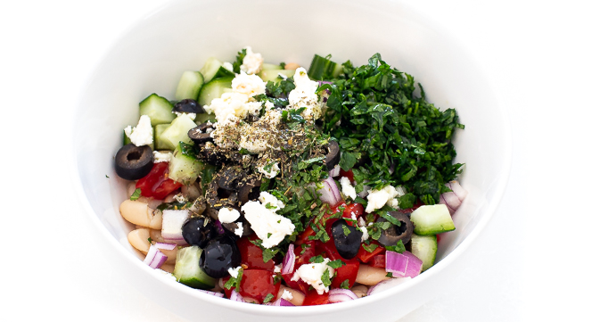 All ingredients for a white bean salad in a white mixing bowl before being mixed.
